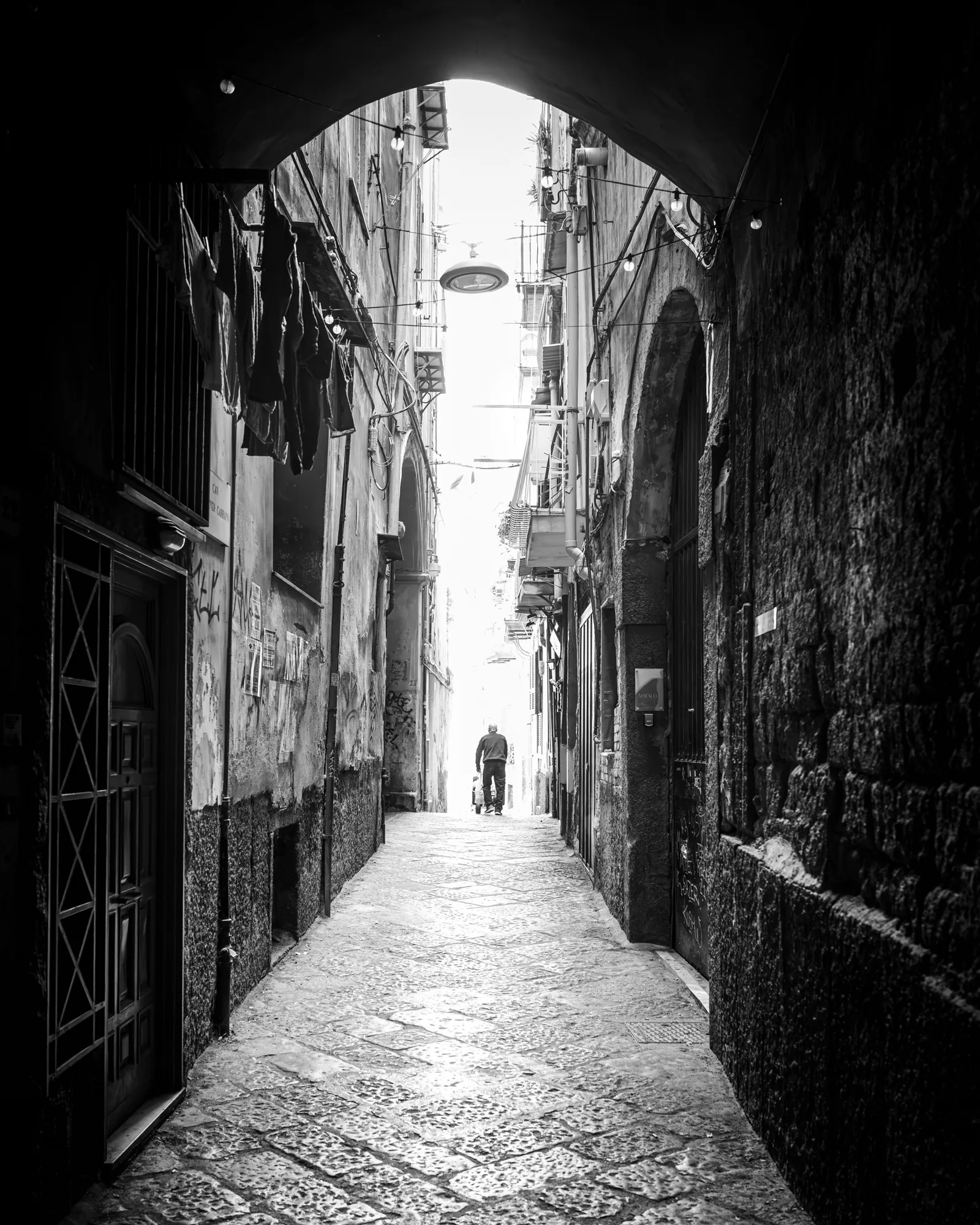 A lone figure at the end of a sunlit alley, framed by a stone archway